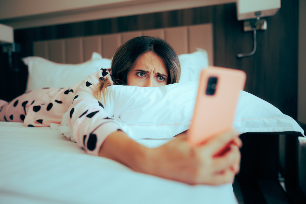 A woman lying on a bed in a room, looking intently at her smartphone with a concerned expression.