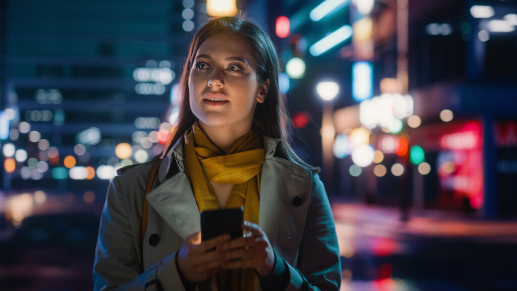  A woman holding a smartphone looks up thoughtfully on a city street at night, with colorful urban lights blurred in the background.