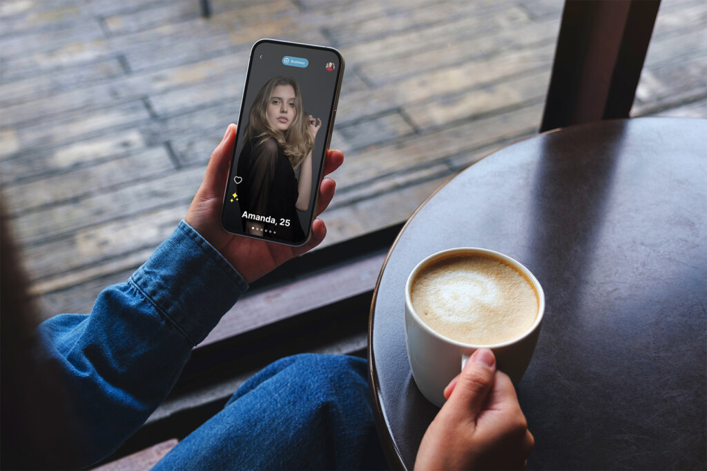 An individual's hand holding a smartphone displaying a dating app profile with a photo of a woman named Amanda, 25, with a cup of coffee on a table in the foreground.