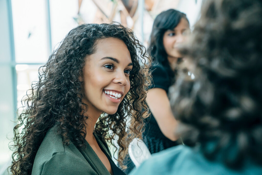 A smiling woman with curly hair, wearing a green blazer, engaging in conversation with someone out of frame.